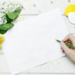 花が置いてある机の上で白紙に何か書こうとしている手の画像