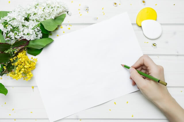 花が置いてある机の上で白紙に何か書こうとしている手の画像